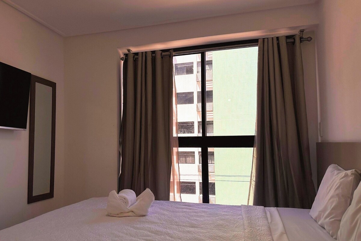 A hotel room with a bed, a flat-screen TV mounted on the wall, and a window with curtains.
