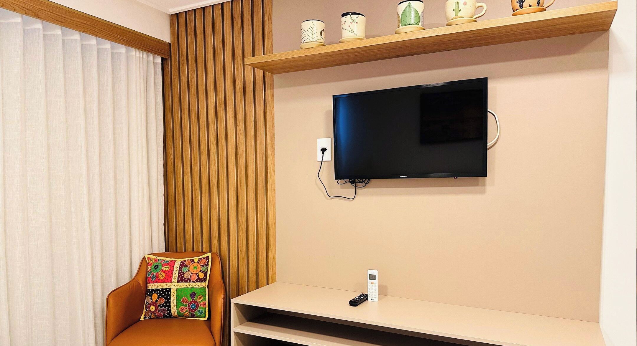 Una habitación con una televisión montada en la pared, un estante de madera encima y una silla con cojín colorido.