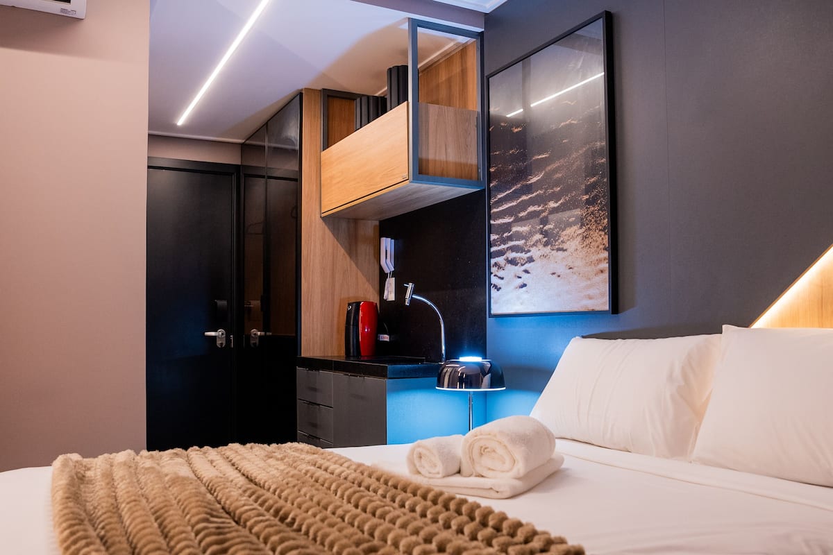 A modern hotel room with a neatly made bed, a wooden cabinet, a desk with a computer, and a large window with a view of the city.