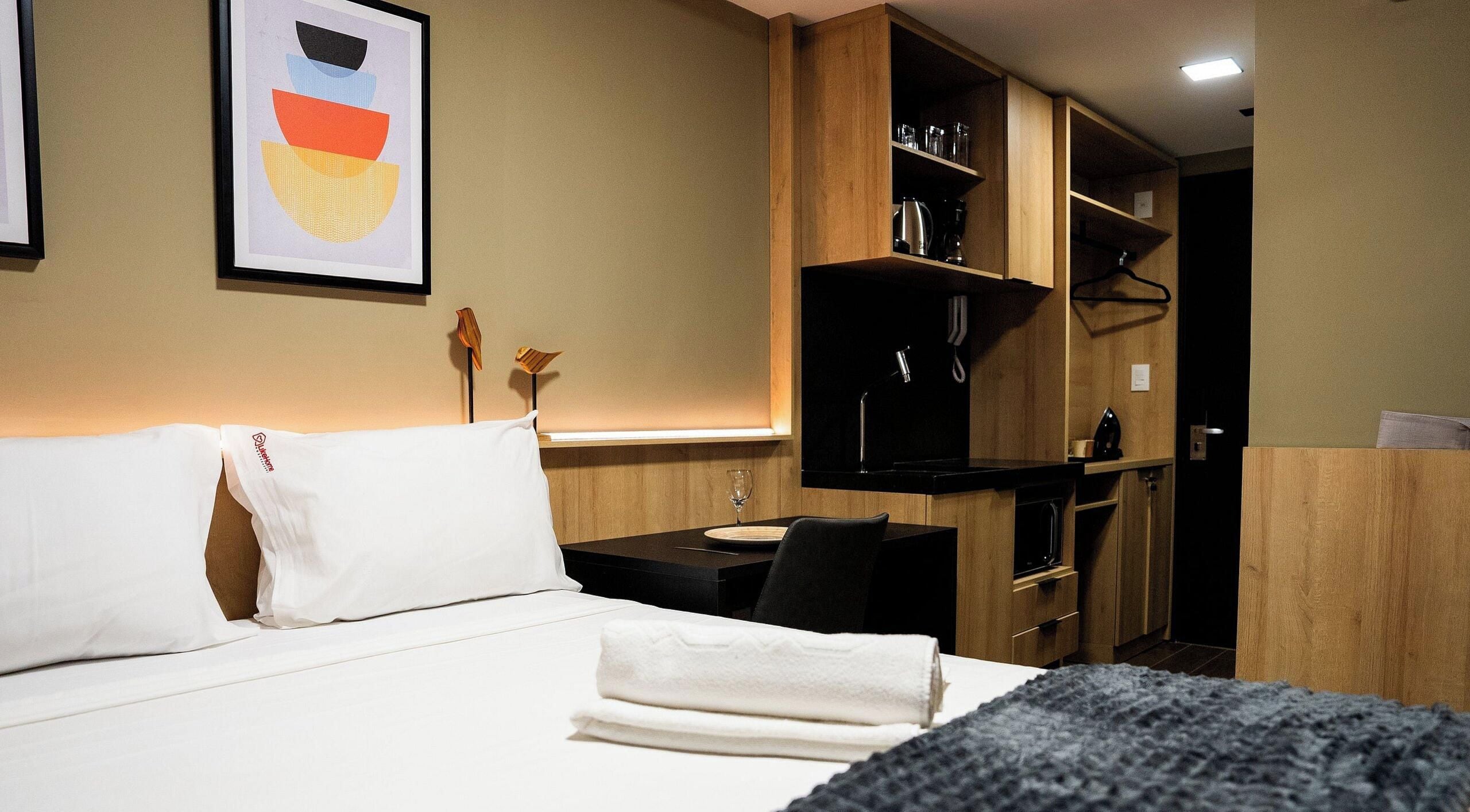 A hotel room with a bed, a desk, a chair, and a kitchenette area.