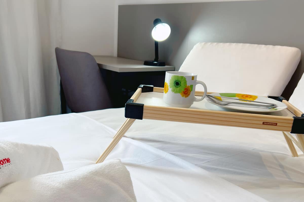 A neatly made bed with a tray holding a mug and chopsticks, a pillow with a logo, and a desk with a lamp in the background.