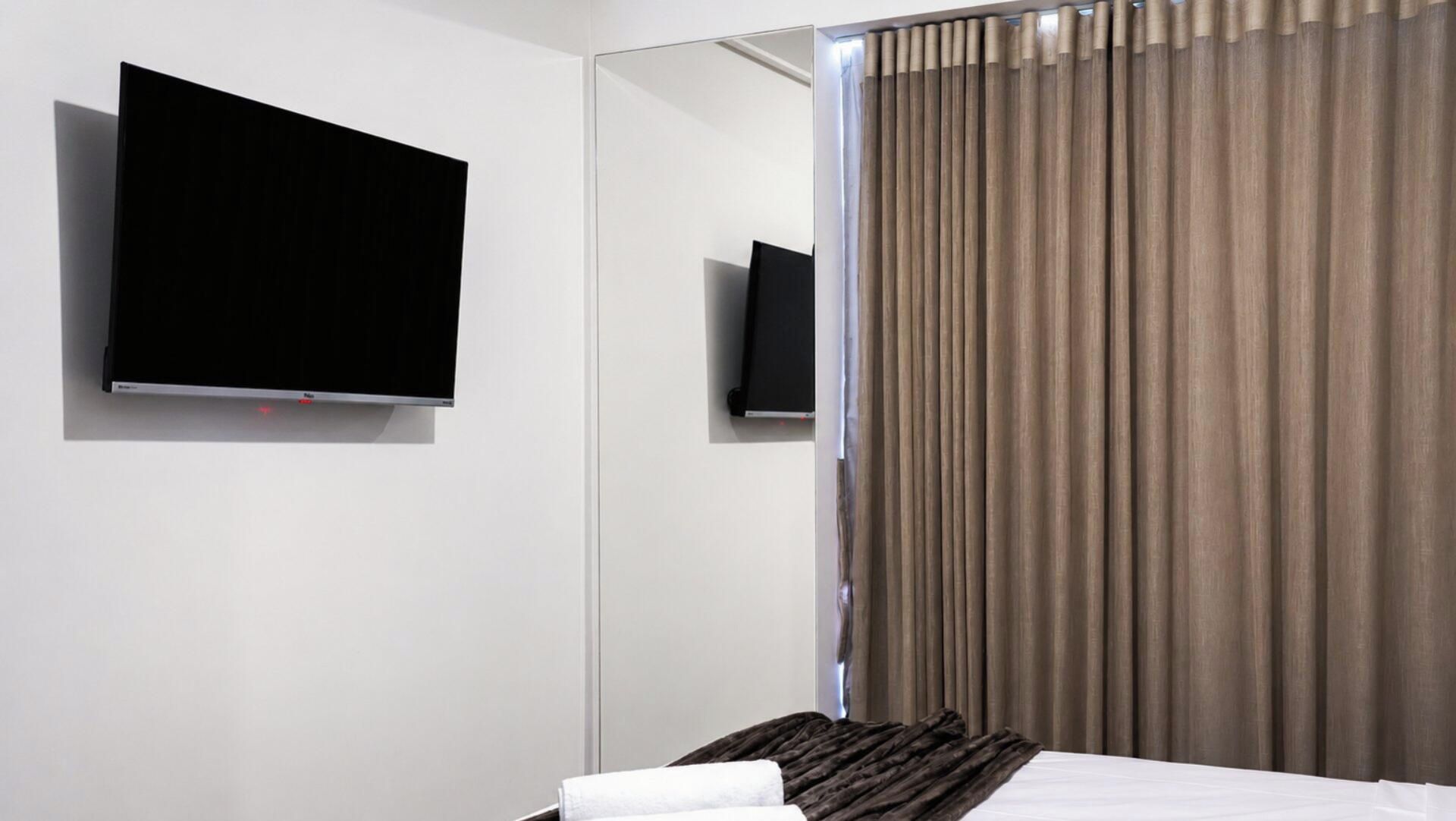 Quarto de hotel com cama, televisão de tela plana na parede e janela com persianas verticais.
