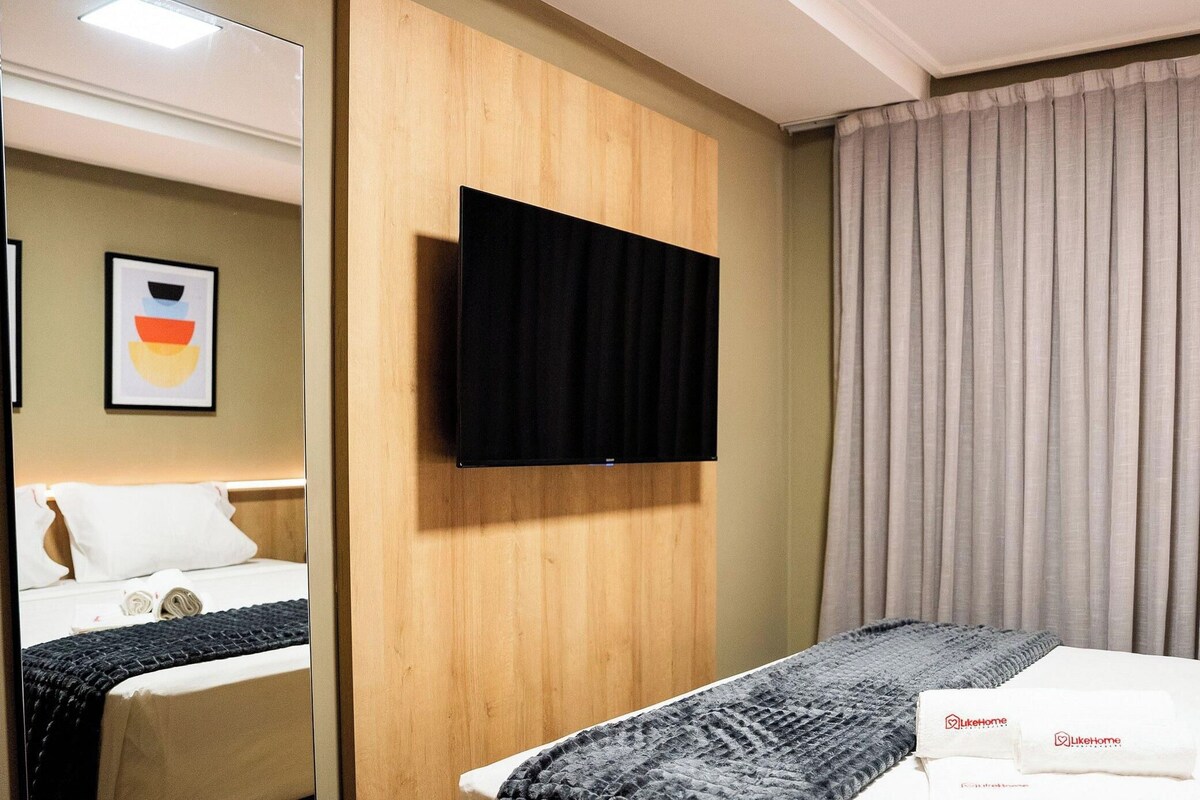 A hotel room with a bed, a flat-screen TV mounted on the wall, a window with curtains, and a framed picture on the wall.