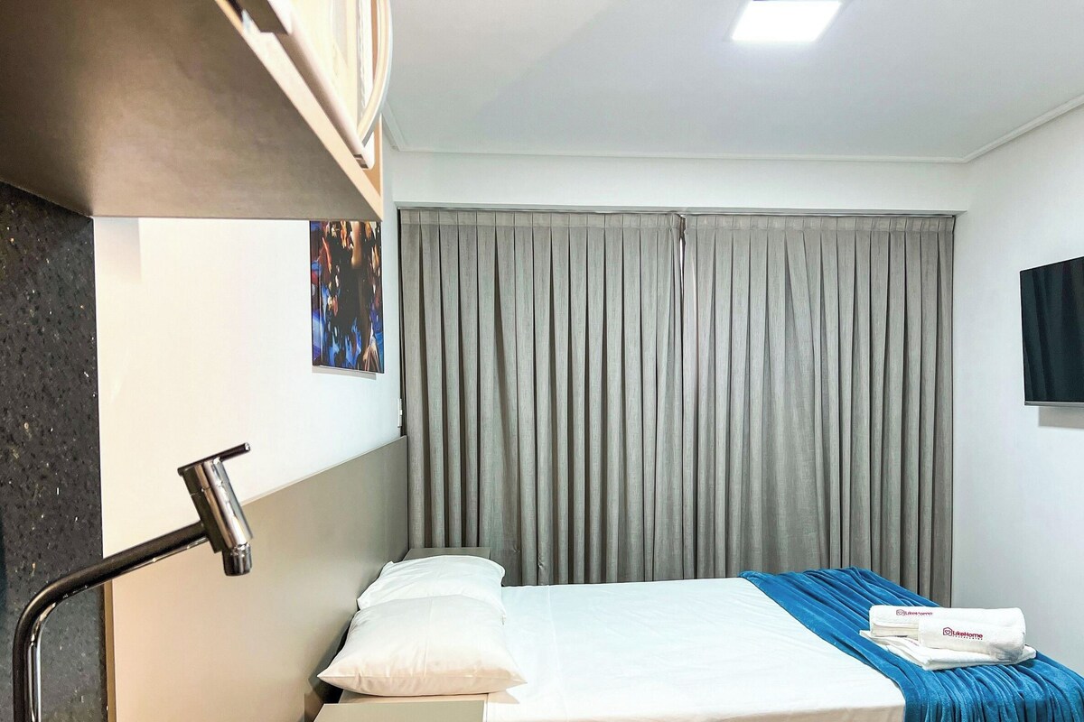 A hotel room with a bed, a flat-screen TV, a window with grey curtains, and a wall-mounted towel dispenser.