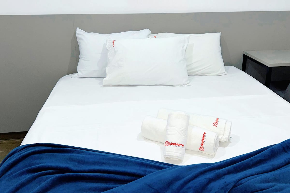 A neatly made bed with white bedding and pillows, a blue blanket, and rolled towels with a logo.