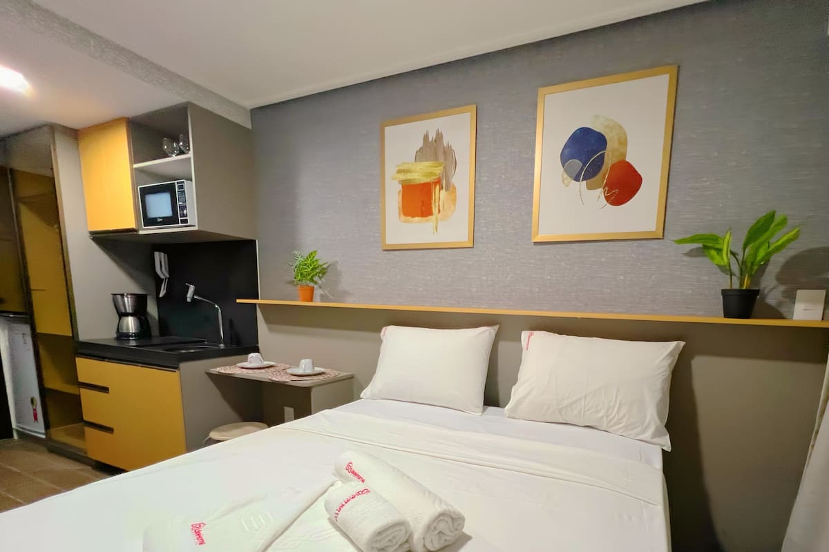 A hotel room with a bed, two pillows, a bedside table with a lamp, a small potted plant, and artwork on the wall.