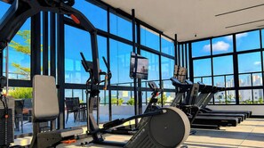A gym with exercise equipment, large windows, and a view of the cityscape. - Move Tambaú (João Pessoa)
