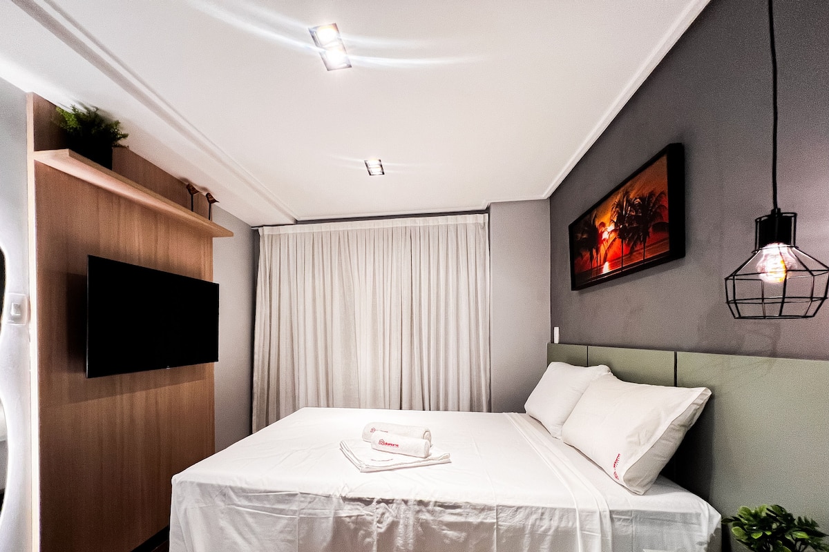 A modern hotel room with a flat-screen TV mounted on the wall, a bed with white bedding, a headboard, and a painting on the wall.