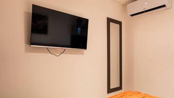 A television mounted on a wall with a black frame, an air conditioning unit above it, and a mirror to the right.