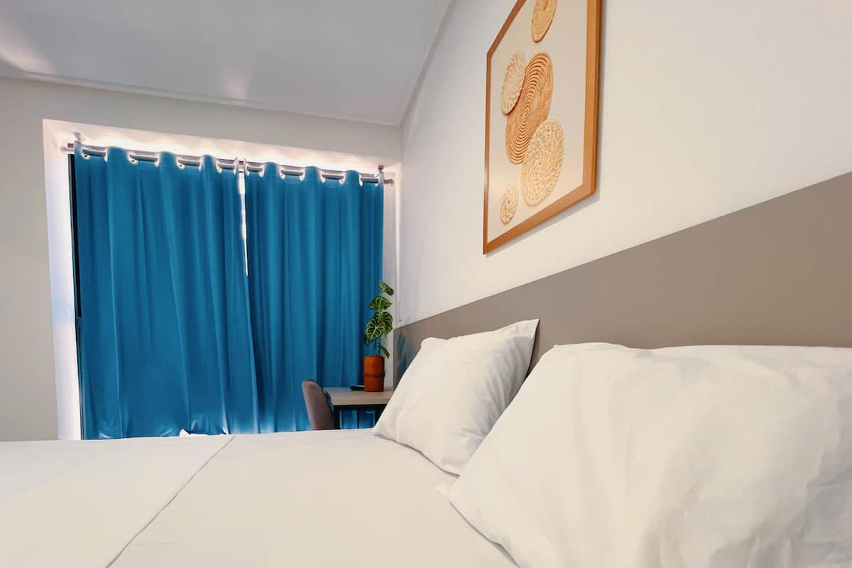 A bedroom with a bed, a window with blue curtains, a small table with a plant, and a framed picture on the wall.