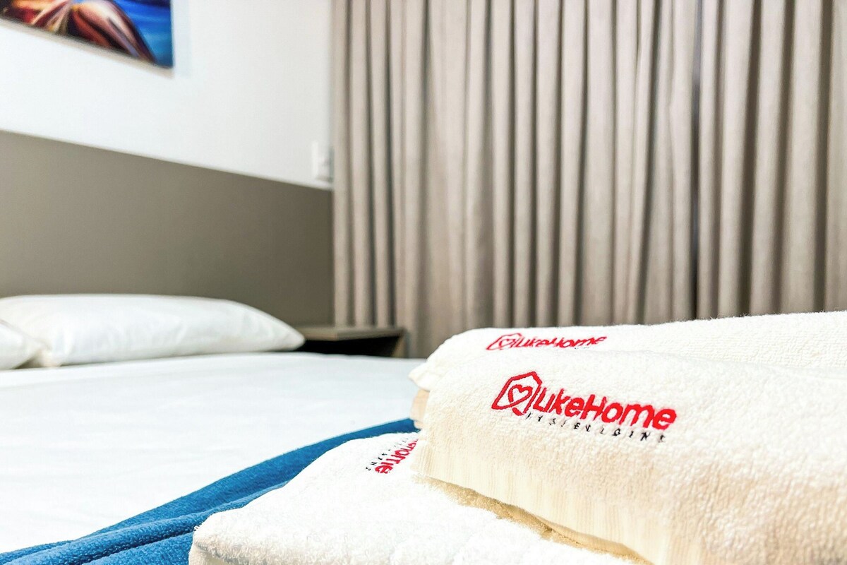 A hotel room with a neatly made bed, a white pillow, and a blue towel. The bed has a white headboard and a blue blanket. The room has a window with curtains.