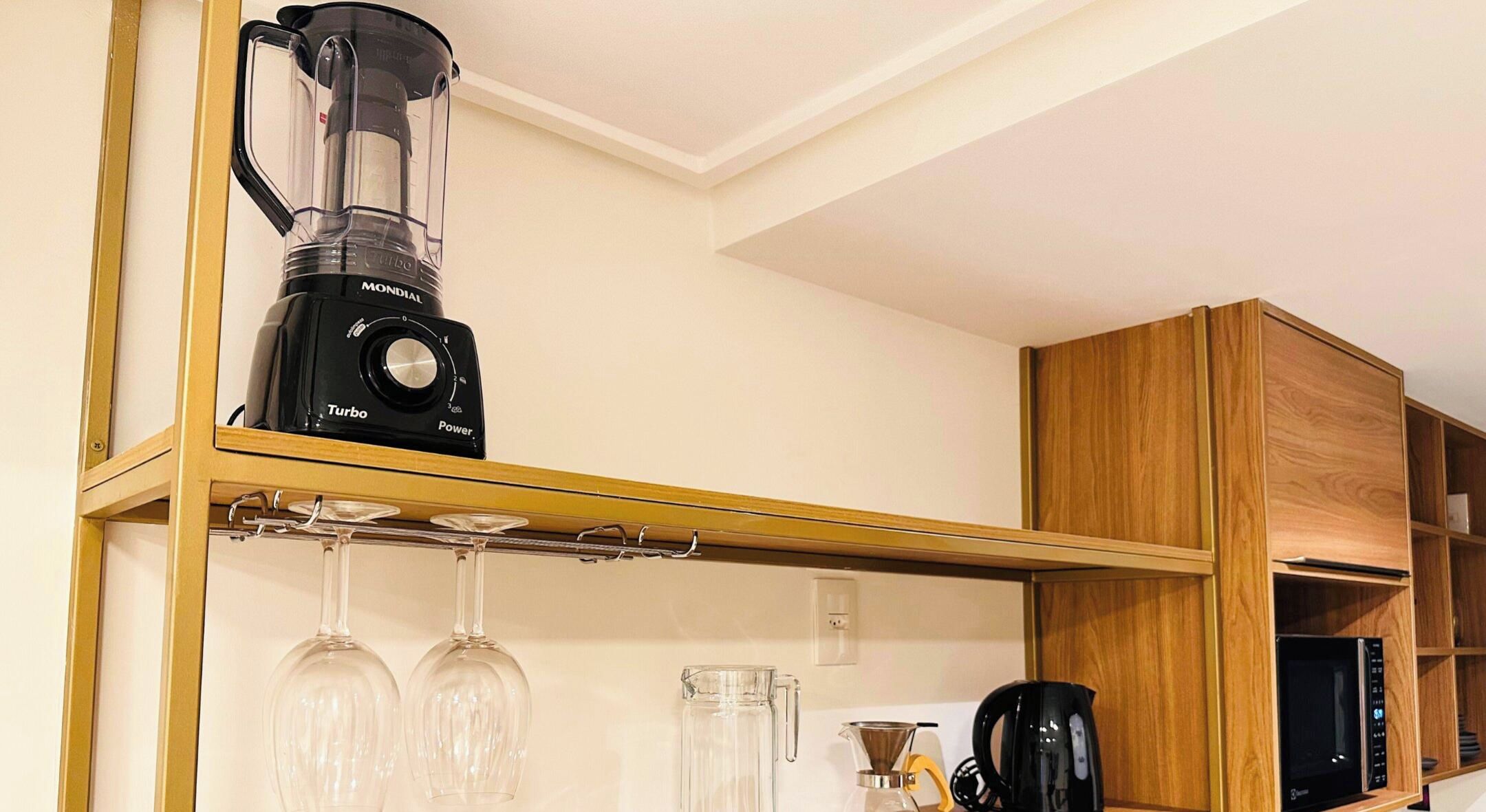 A kitchen with a blender on a shelf, wine glasses, a coffee maker, and wooden cabinets.