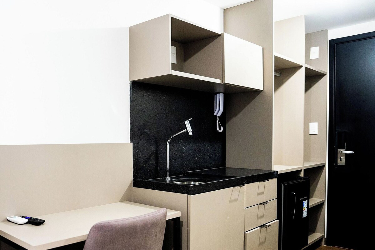 A compact kitchenette with a black countertop, a faucet, and a small sink. There is a white desk with a remote control on it.