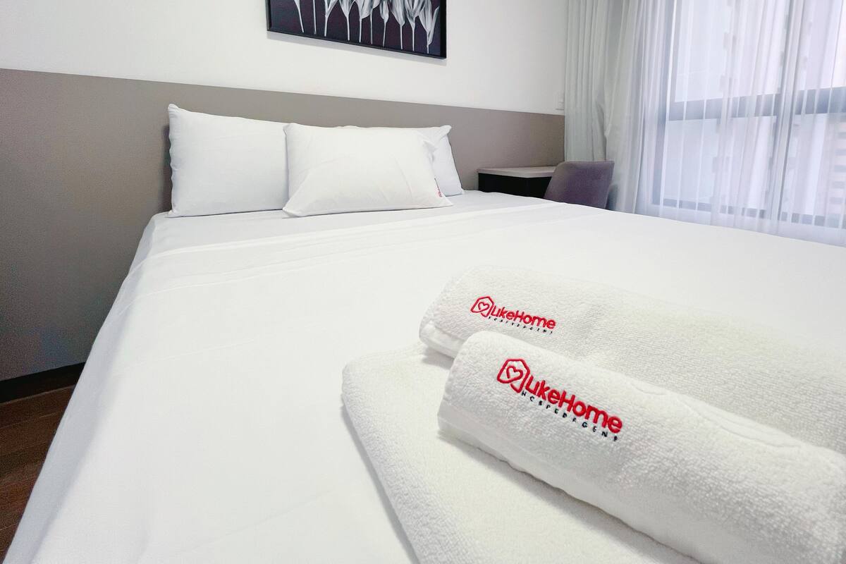A neatly made bed with white linens and two towels with 'LikeHome' branding.