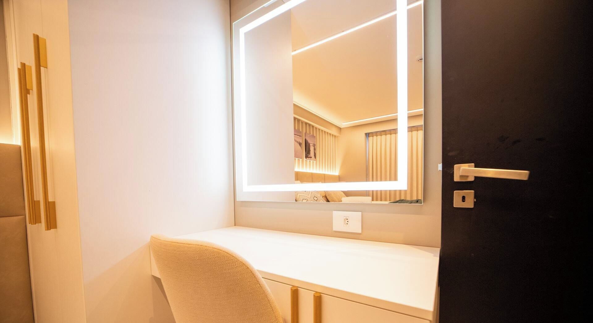 A hotel room with a bedside table, a chair, a mirror with integrated lighting, a door with a handle, and a wall socket.