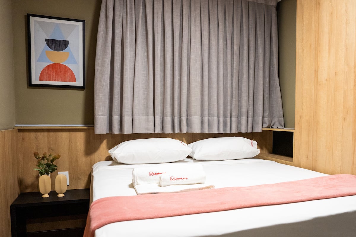A neatly made bed with white bedding, a pink throw, and a set of white pillows. There is a framed artwork on the wall above the bed, and a wooden nightstand with a small plant and a glass of water beside it.