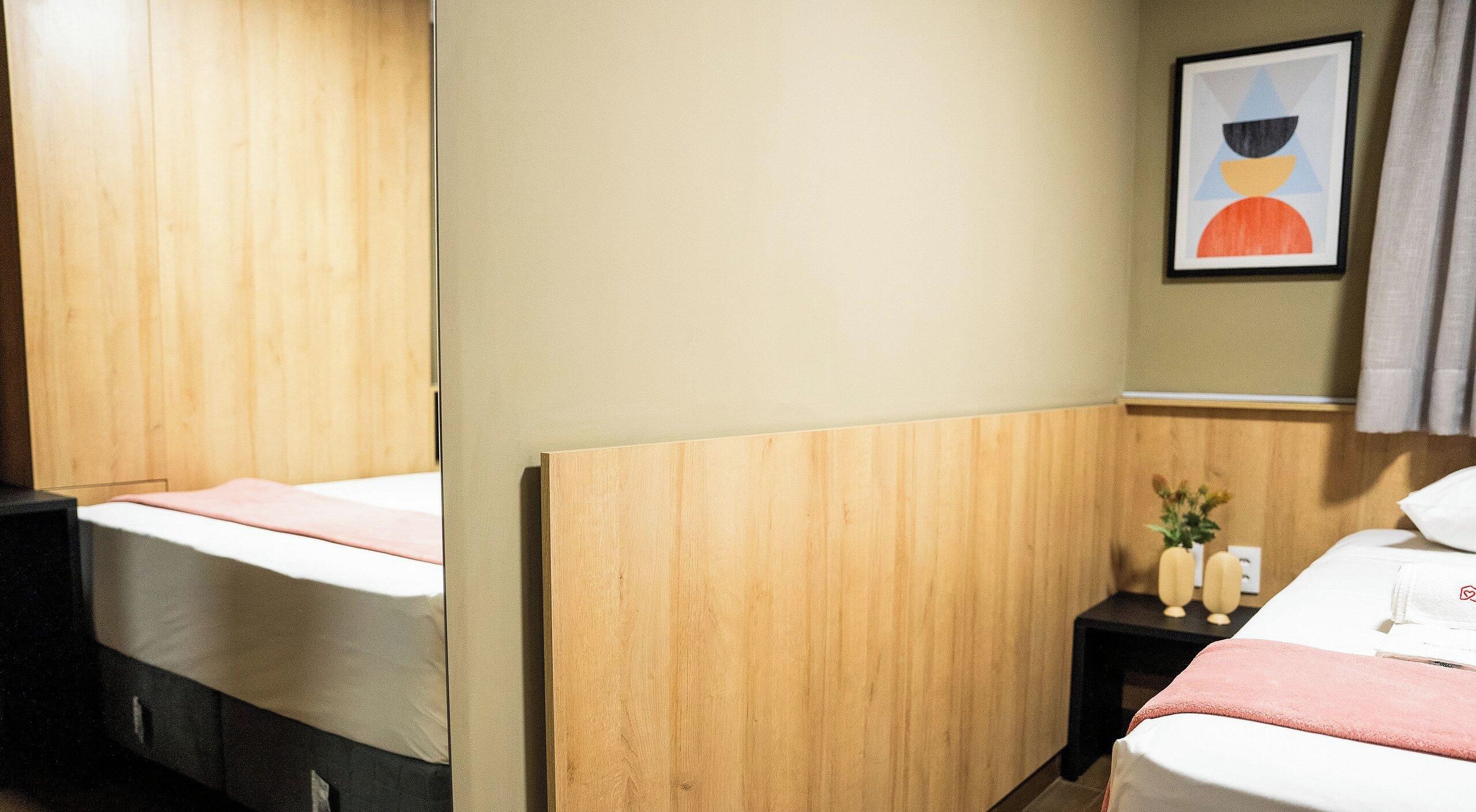 Una habitación de hotel con cabecera de madera, cama con colchón blanco, mesita de noche con un florero y una lámpara, y un cuadro en la pared.