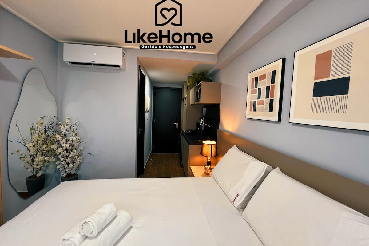 A hotel room with a bed, a bedside lamp, a wall-mounted air conditioner, a potted plant, and framed artwork.