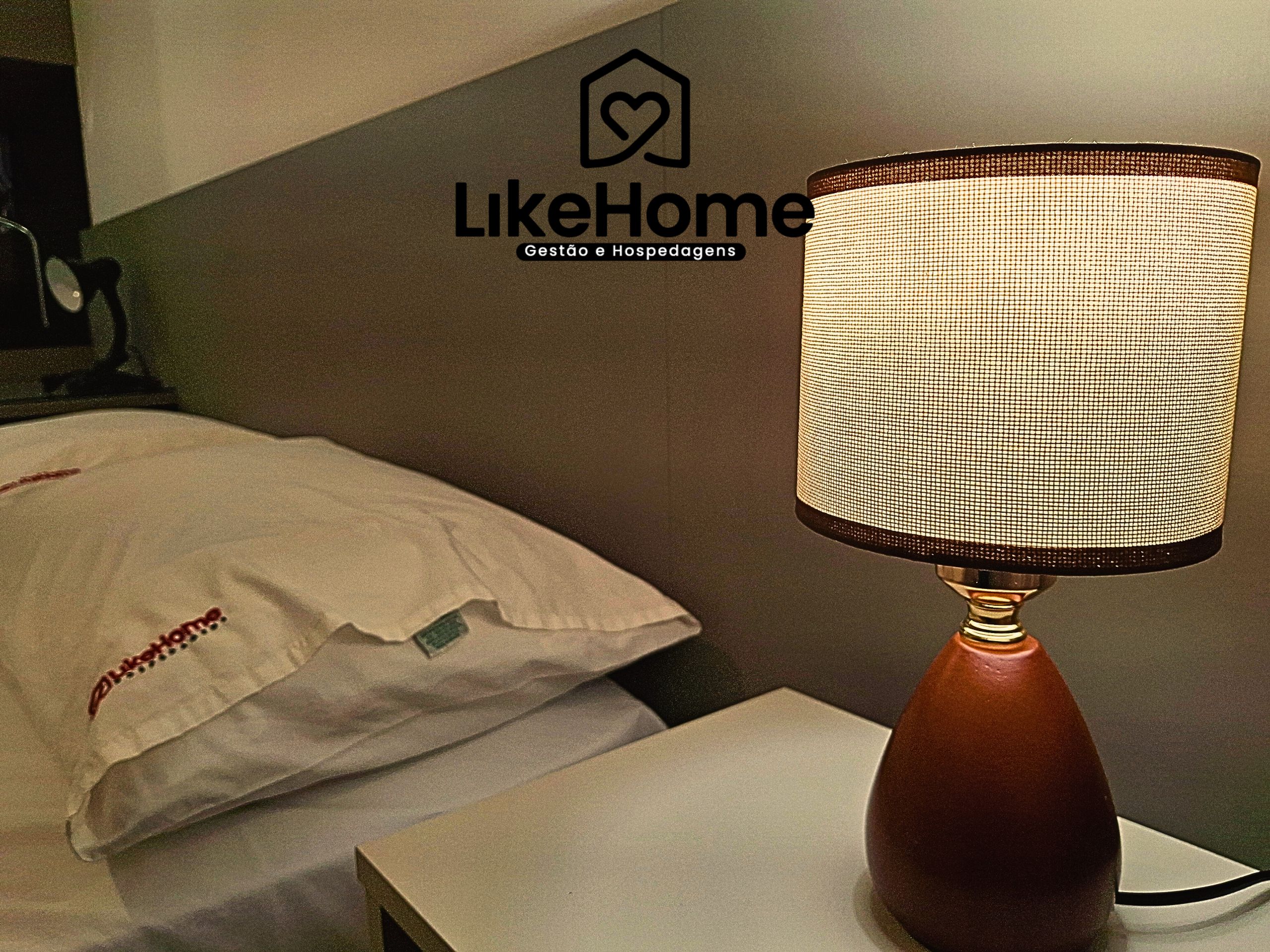 Egy szállodai szoba, amelyben található egy ágy, egy lámpa és egy fal, amelyen a LikeHome logó látható.