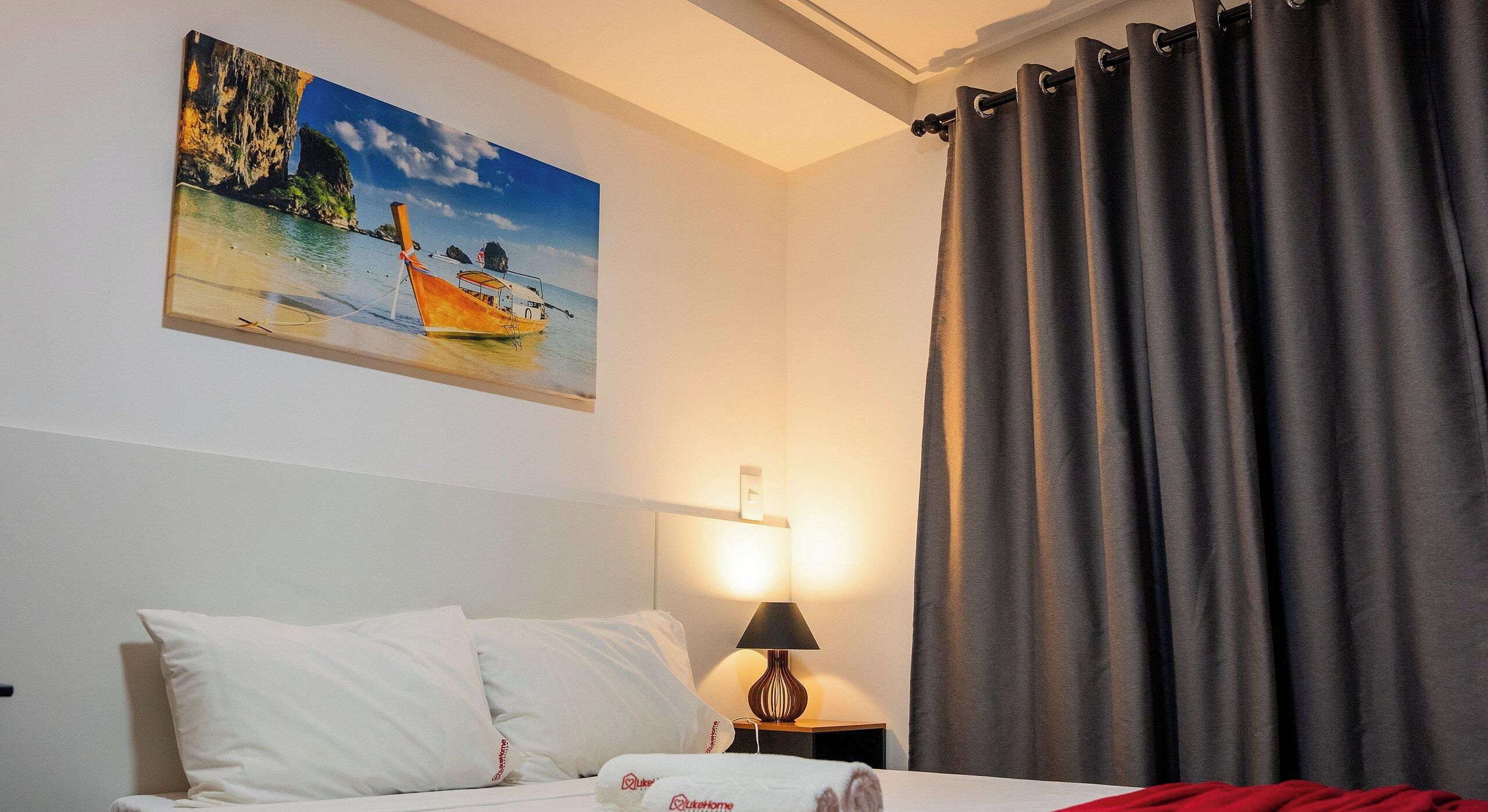 A hotel room with a bed, a painting of a coastal scene, a lamp, and grey curtains.