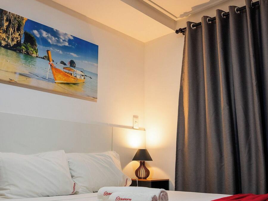 Quarto de hotel com uma cama, um quadro de uma paisagem costeira, um abajur e cortinas cinzentas.