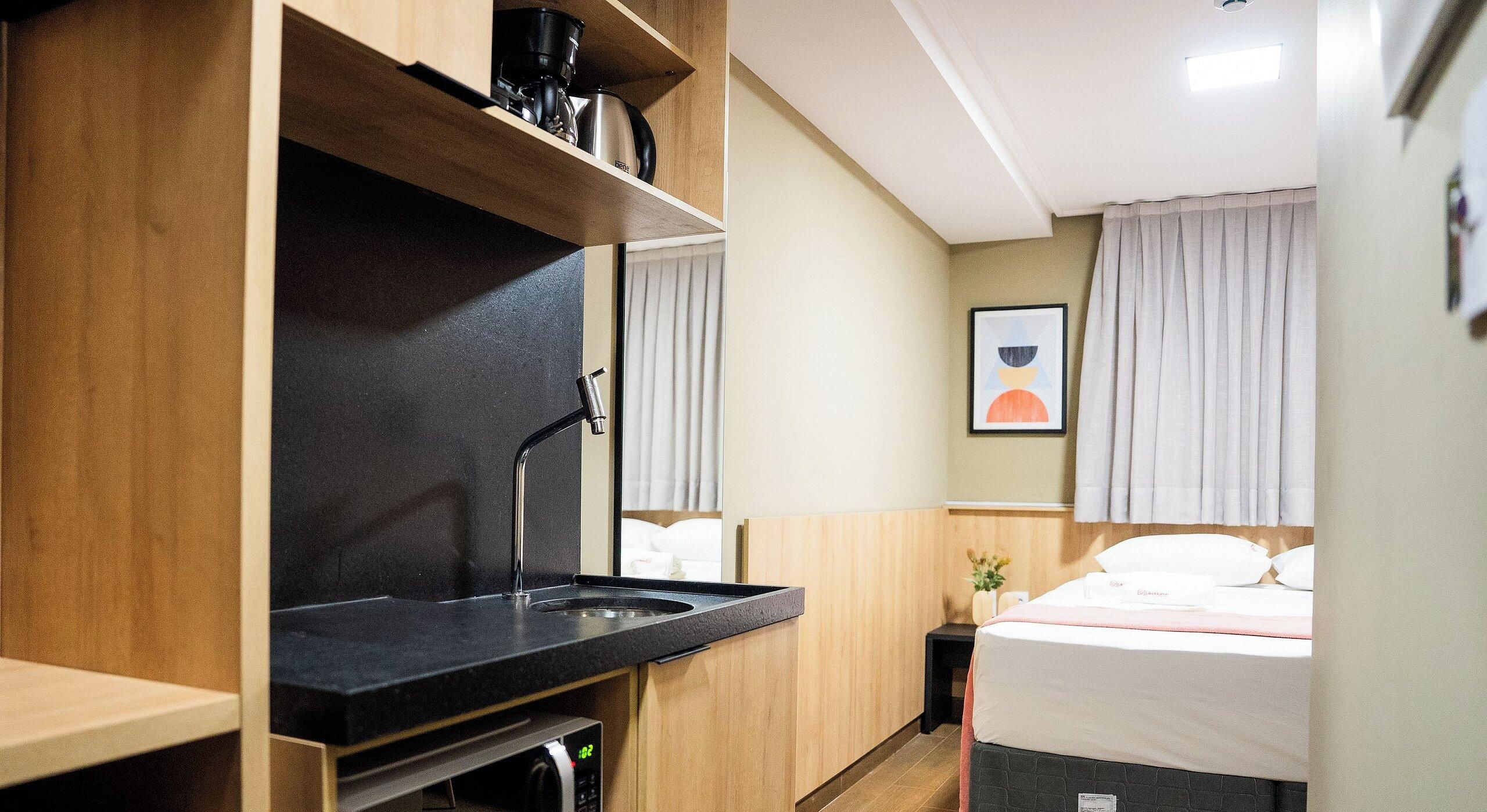 Habitación de hotel compacta con kitchenette, una cama con ropa blanca, una mesita de noche pequeña y una obra de arte colgada en la pared.