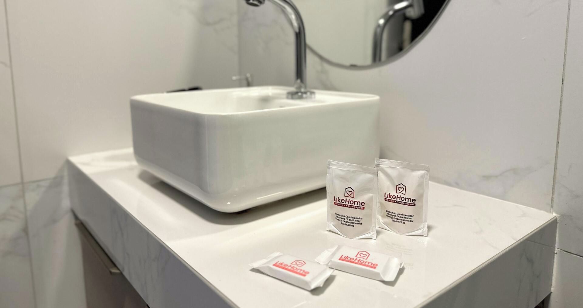 Une salle de bain avec un lavabo blanc et deux échantillons de shampoing de la marque LikeHome posés sur le comptoir.