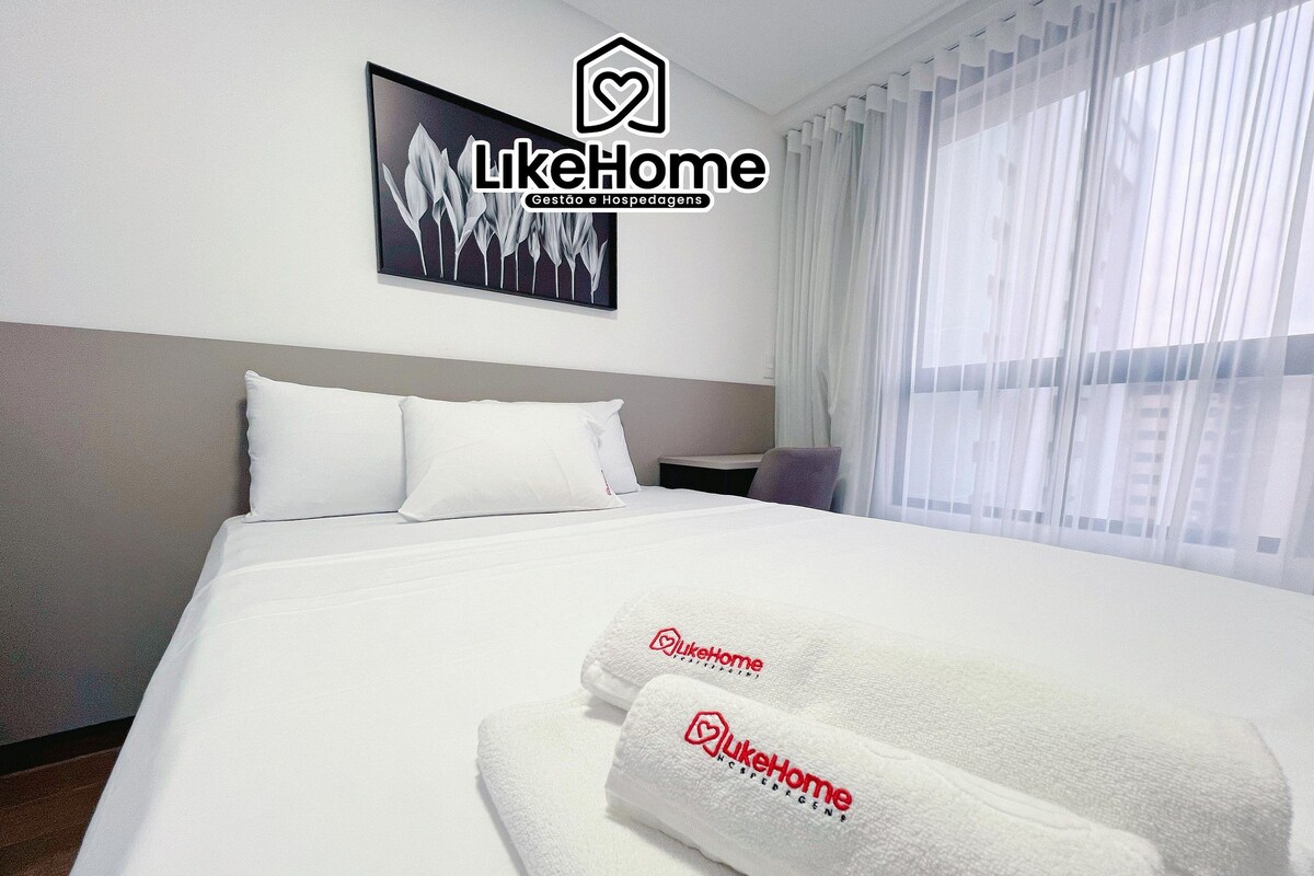 A hotel room with a neatly made bed, a headboard, a bedside table, and a window with sheer curtains.