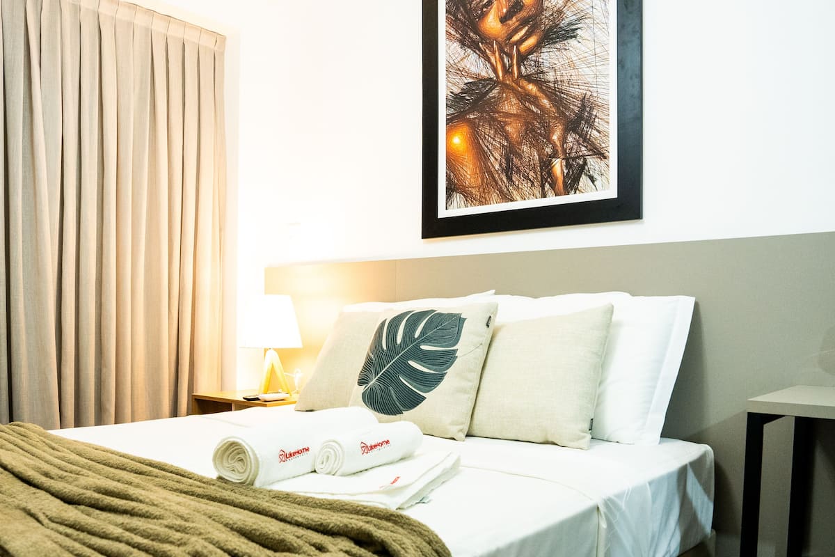 A hotel room with a bed, a large window with curtains, a nightstand with a lamp, and a framed artwork on the wall.