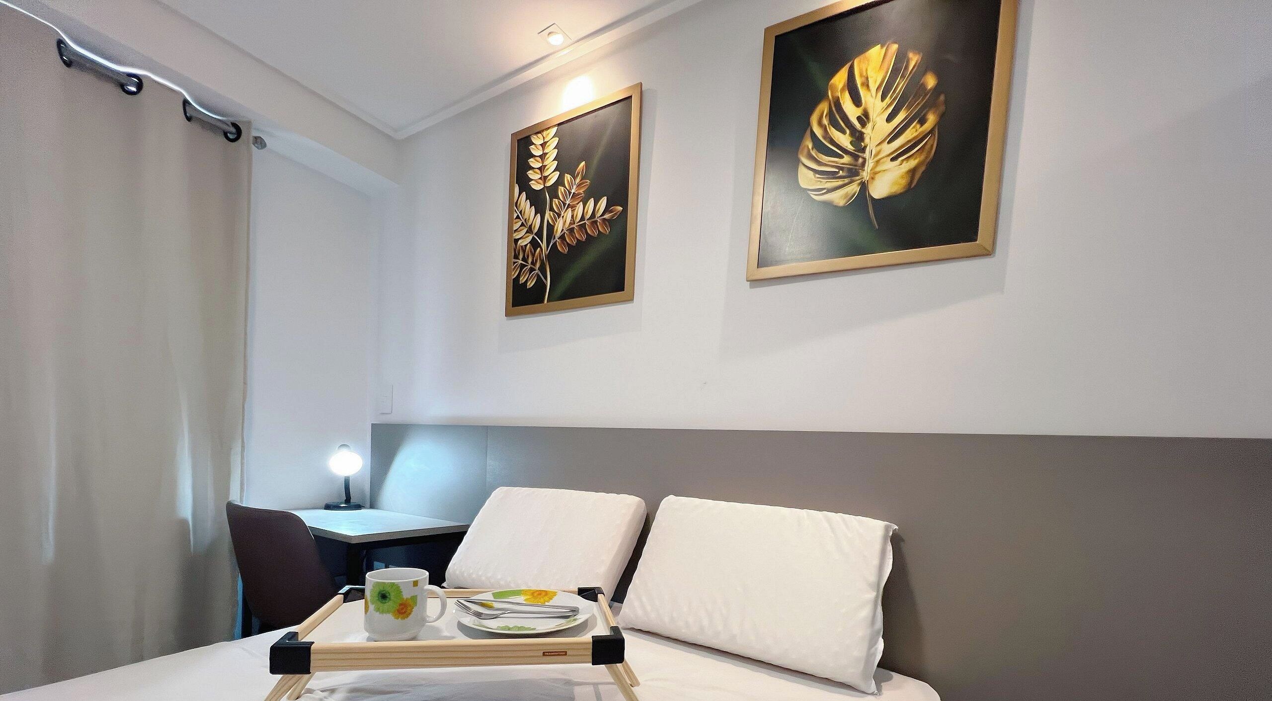 A hotel room with a bed, a desk, and two framed artworks on the wall.