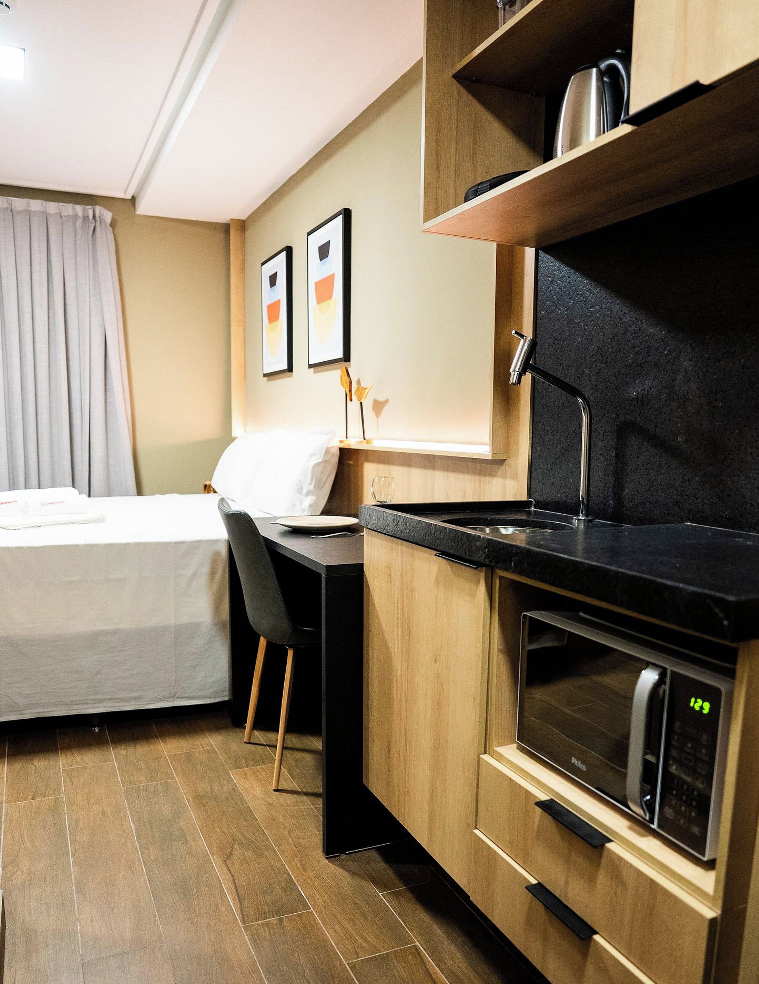 A compact kitchenette with a microwave, a small oven, and a countertop. There is a bed with a white pillow and a chair.