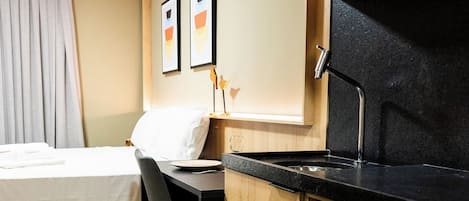 A compact kitchenette with a microwave, a small oven, and a countertop. There is a bed with a white pillow and a chair.