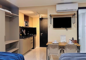 Ett kompakt hotellrum med ett litet pentry, en matplats med bord och stolar, en TV som är monterad på väggen och en säng med ett blått täcke.