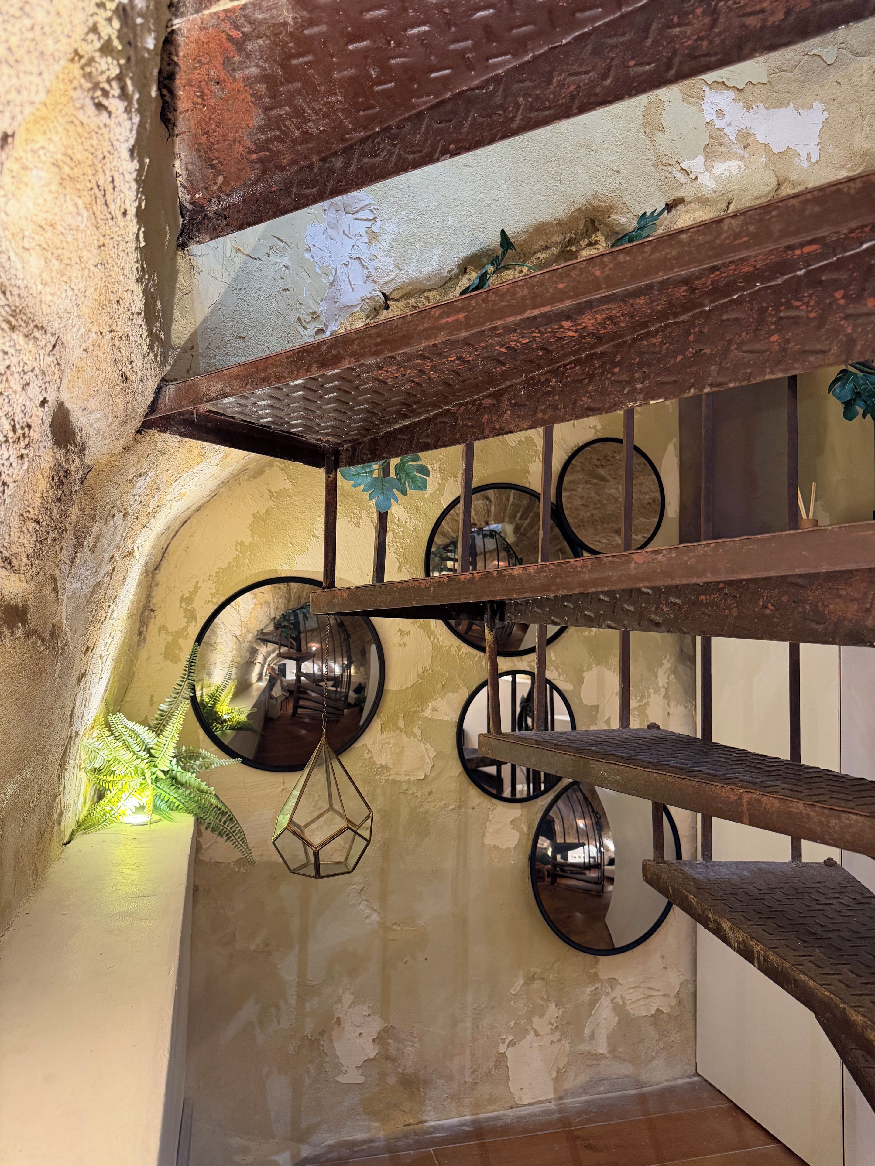 Un intérieur rustique avec des murs en briques apparentes, un escalier en métal et un grand miroir rond accroché au mur.