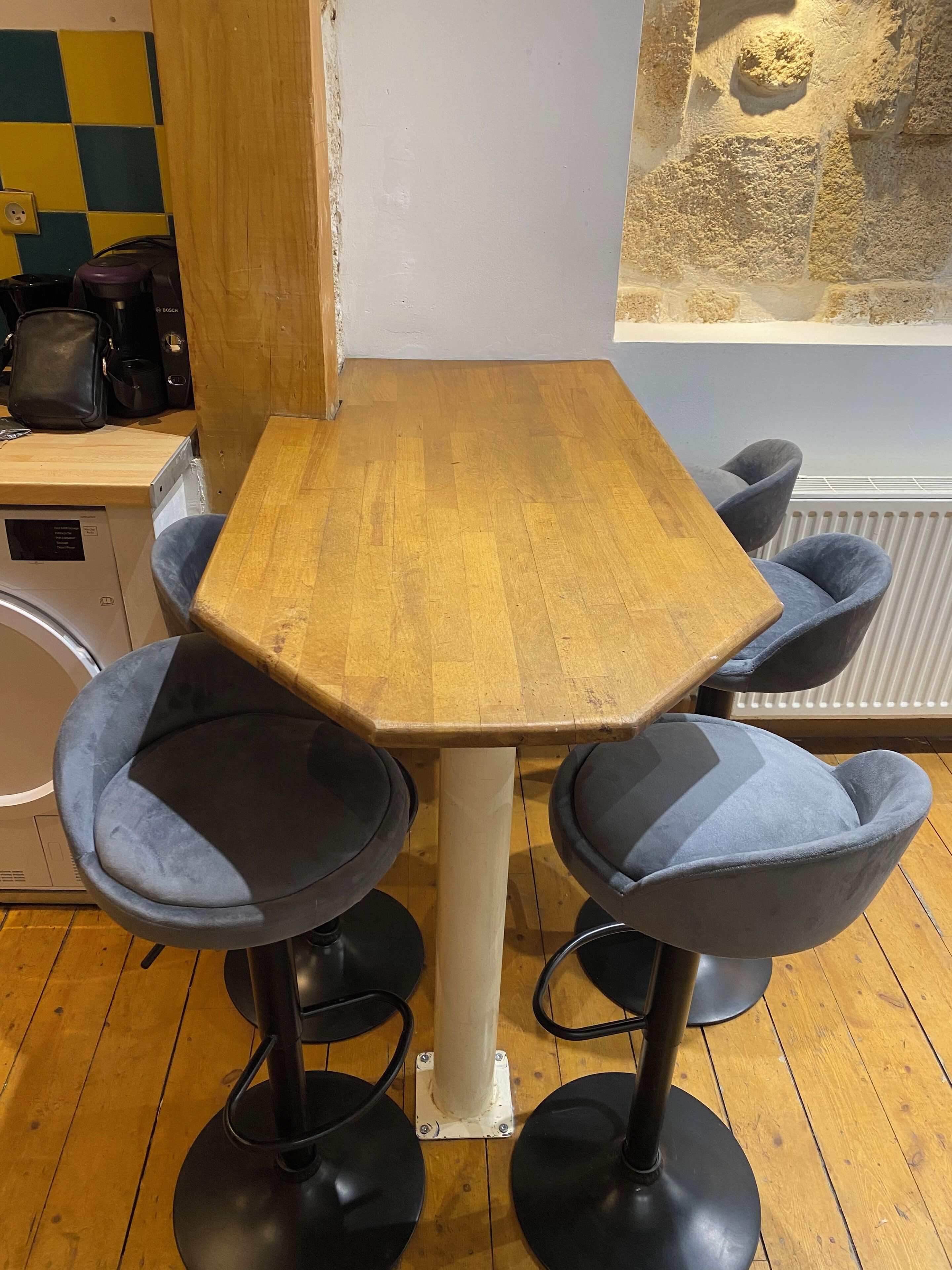 Une table en bois, quatre chaises bleues, une machine à café et un radiateur à l’arrière-plan.