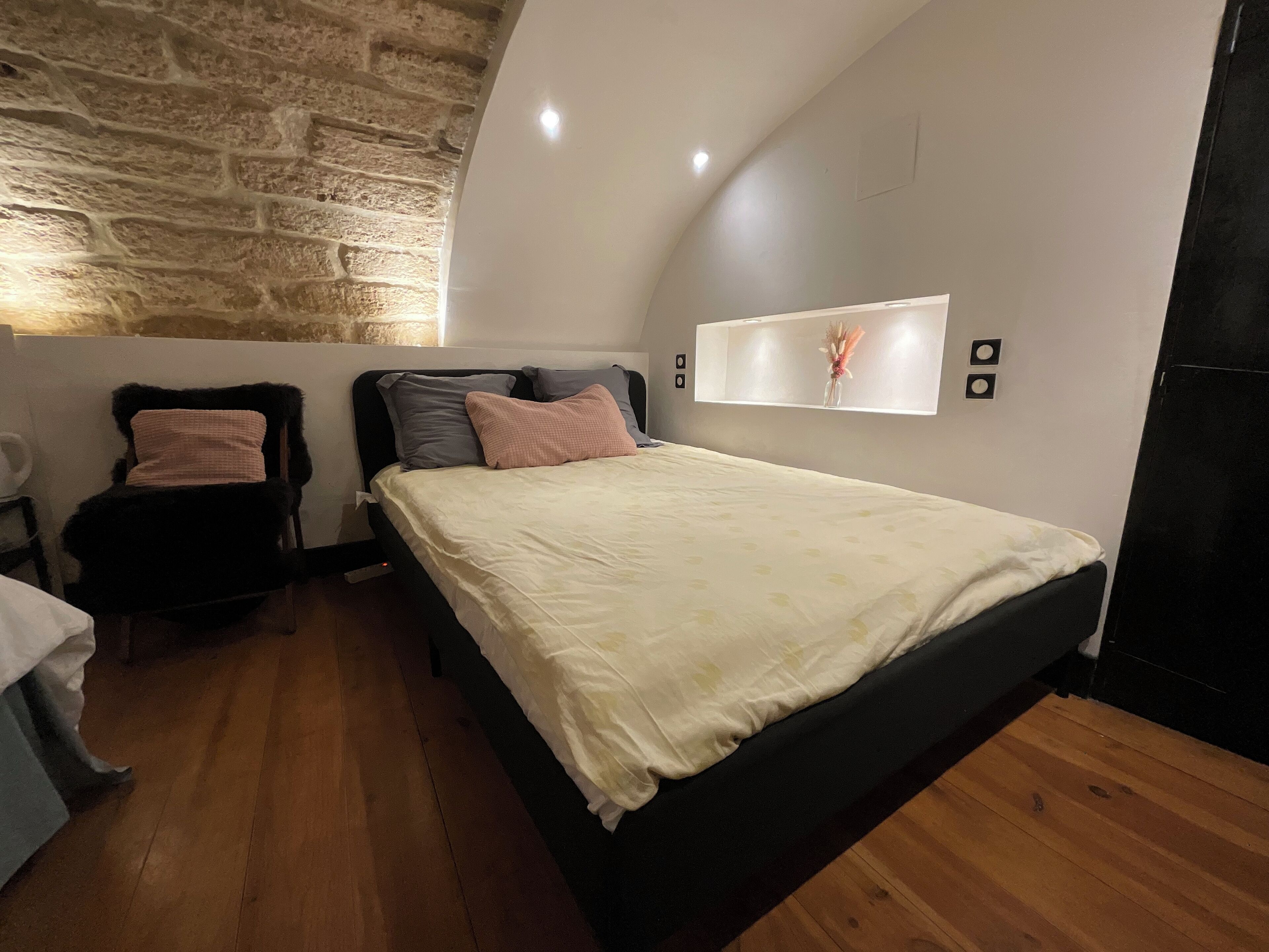 Une chambre avec un mur en pierre, un lit recouvert d’une couette blanche, une chaise agrémentée d’un plaid en fausse fourrure noire et une table de chevet avec une lampe.