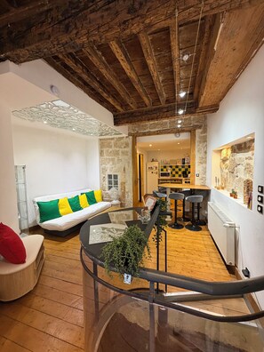 Un salon chaleureux avec un plafond en bois, une table basse en verre agrémentée d’une plante, un canapé avec des coussins colorés et, au fond, un coin cuisine.