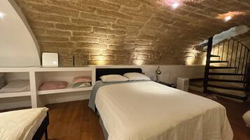 Une chambre avec un plafond en pierre, un lit avec une literie blanche, une table de chevet, une lampe et une petite étagère avec des sacs.