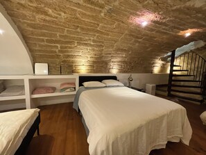 A bedroom with a stone ceiling, a bed with white bedding, a bedside table, a lamp, and a small shelf with bags.
