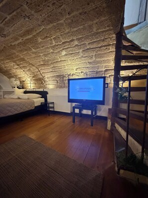 Une chambre mansardée chaleureuse, avec un téléviseur posé sur un meuble en bois, un lit recouvert d’une courtepointe, un radiateur et un escalier en bois.