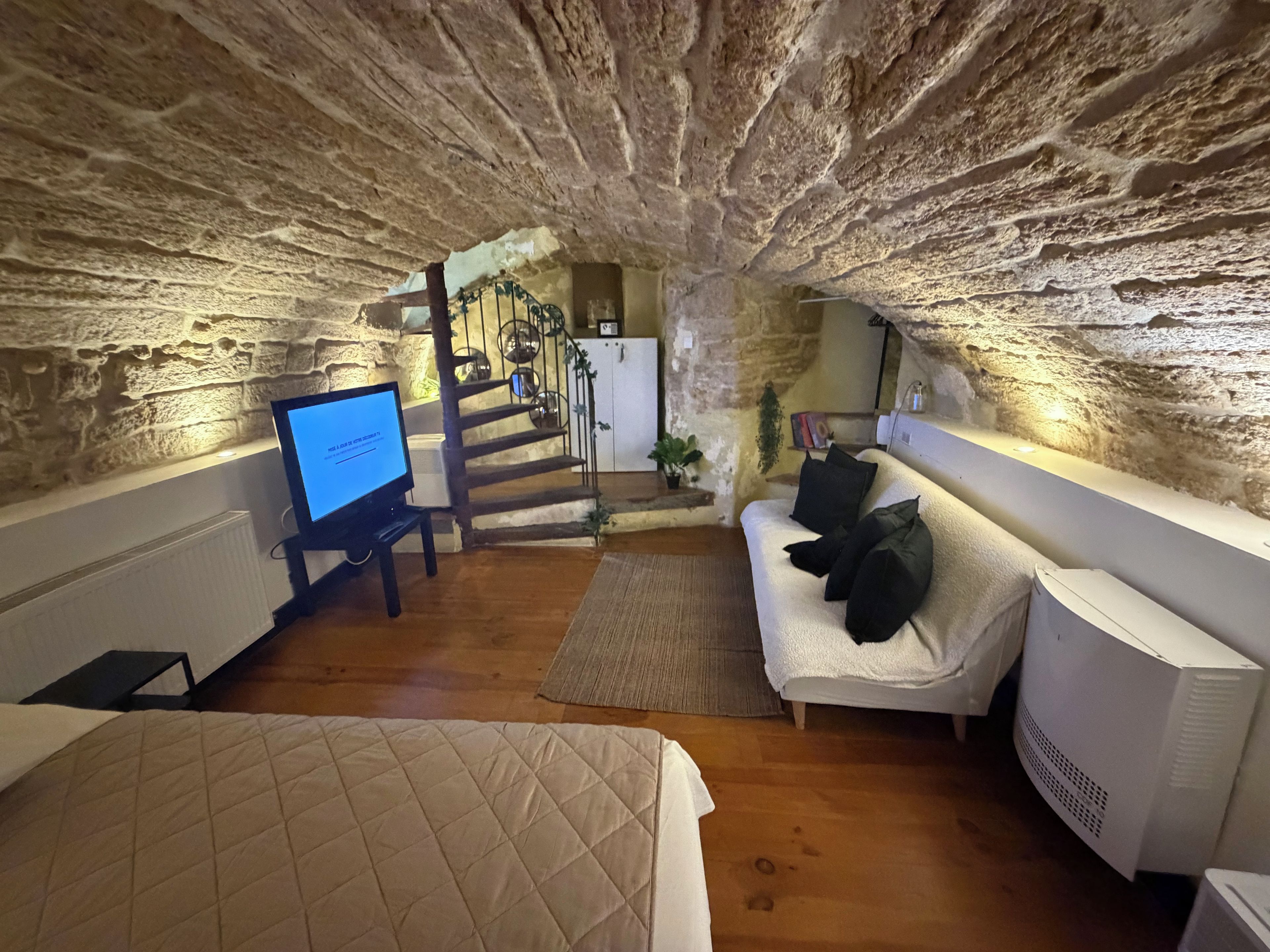 Ein gemütliches Zimmer mit Steinwand, einem Bett, einer Couch mit Kissen, einem Fernseher und einer Treppe, die in einen Küchenbereich führt.