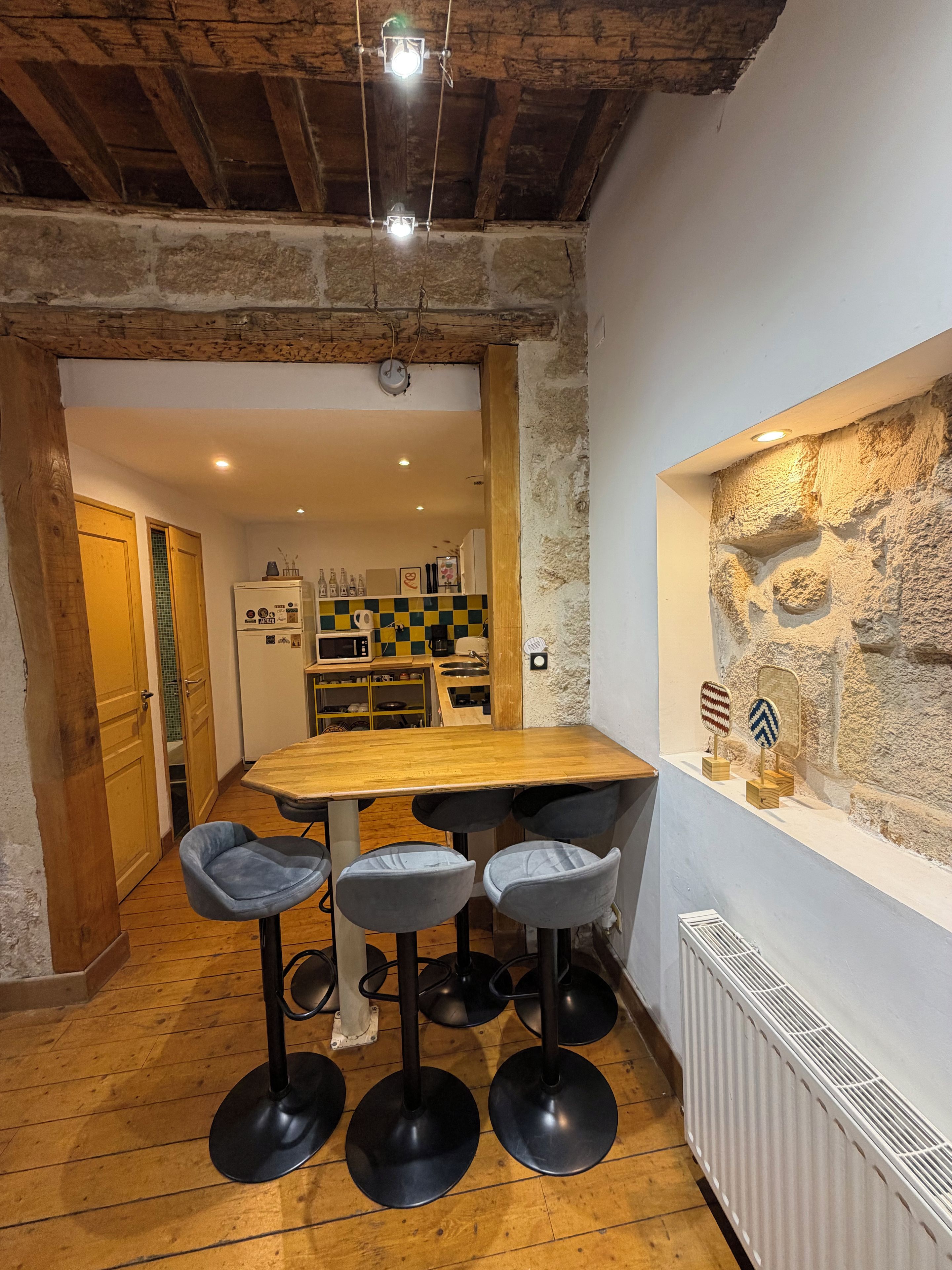 Une cuisine rustique avec une table en bois et des tabourets de bar, un mur en pierre avec une fenêtre, et un comptoir de cuisine apparent avec des appareils électroménagers.