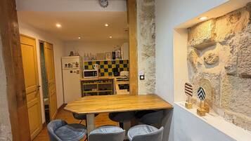 Une cuisine rustique avec une table en bois et des tabourets de bar, un mur en pierre avec une fenêtre, et un comptoir de cuisine apparent avec des appareils électroménagers.