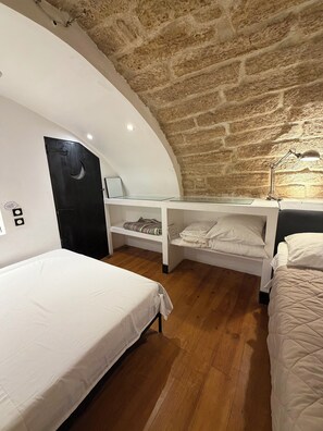 Une chambre à coucher avec un plafond incurvé, un plancher en bois et un mur en pierre. On y trouve deux lits avec du linge de lit blanc, un bureau avec une lampe et une petite étagère.