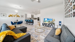 A modern living room with a grey sofa, a coffee table, and a TV. There's a kitchen in the background with a bar area.