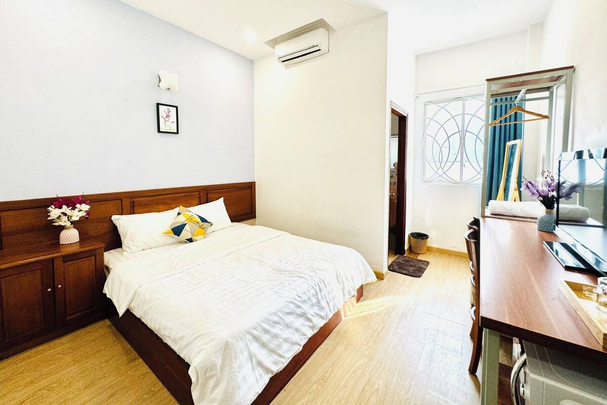 Een hotelkamer met een houten bed, wit beddengoed, een klein nachtkastje met een bloemenvaas, een wandairconditioner en uitzicht op buiten door een raam.