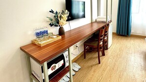 A compact room with a wooden desk, a mounted TV, a wall fan, a small seating area, and a window with curtains. - Huyen Home (Ho Chi Minh City)