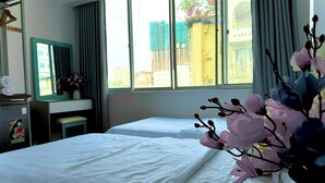 A hotel room with two beds, a desk, a chair, and a large window with a view of buildings. - Huyen Home (Ho Chi Minh City)