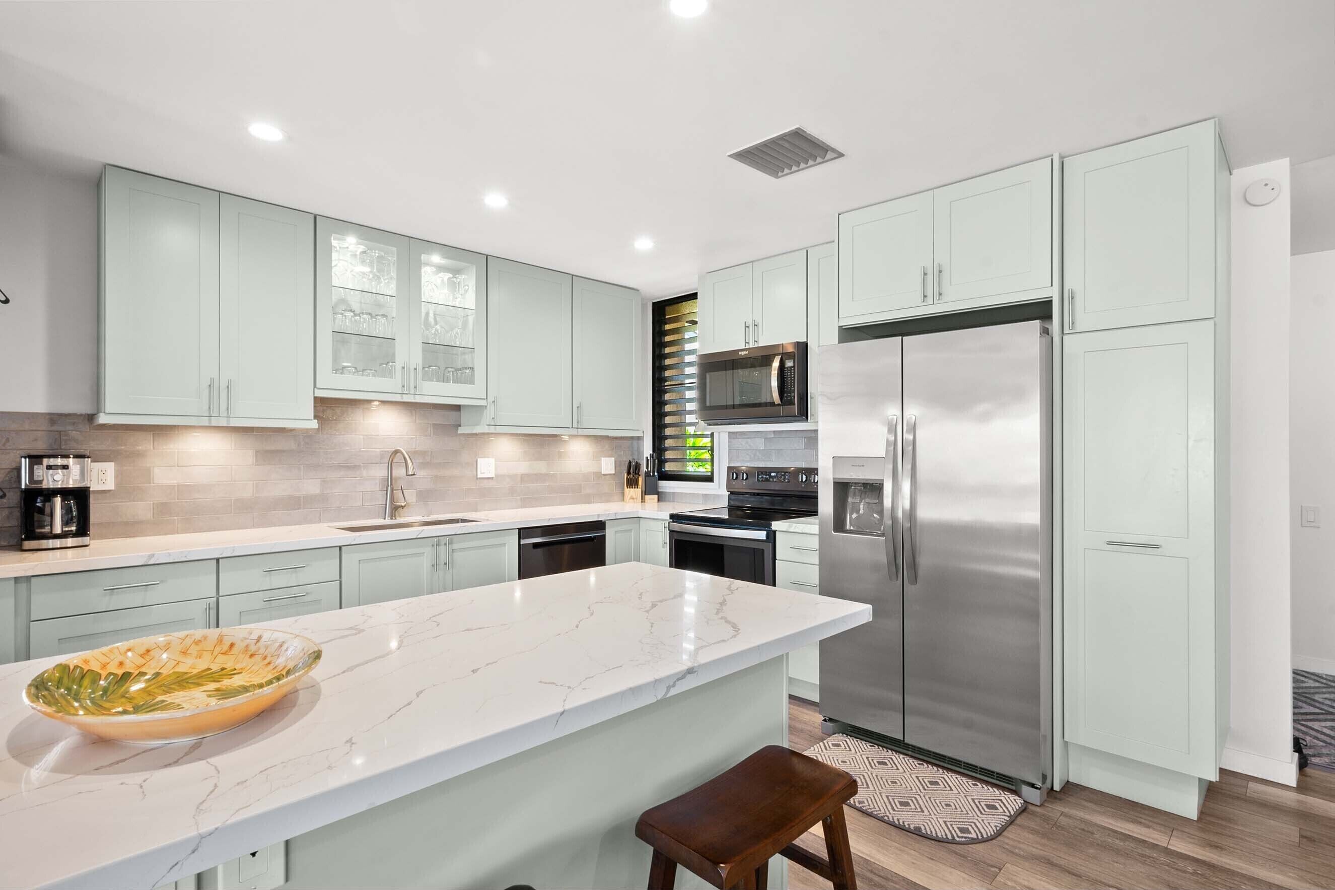 A modern kitchen with light green cabinets, a marble countertop, stainless steel appliances, and a large island with a bowl.