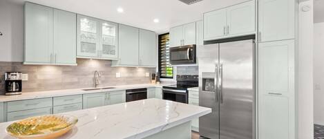 A modern kitchen with light green cabinets, a marble countertop, stainless steel appliances, and a large island with a bowl.
